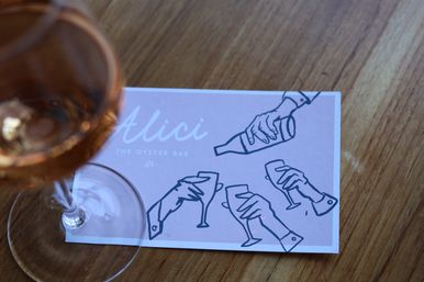Rosé wine glass on a wooden table next to a pink oyster-bar card featuring playful line drawings of hands pouring and toasting.