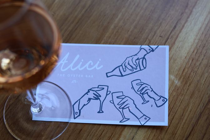 Rosé wine glass on a wooden table next to a pink oyster-bar card featuring playful line drawings of hands pouring and toasting.