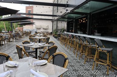 Al fresco bistro patio with black-and-white patterned tile, marble tables set for dining, rattan woven chairs, bar stools, umbrellas and string lights
