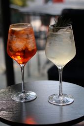Two chilled cocktails in wine glasses on a round outdoor bistro table — an amber spritz with ice and a lemon slice and a pale citrus cocktail garnished with rosemary.