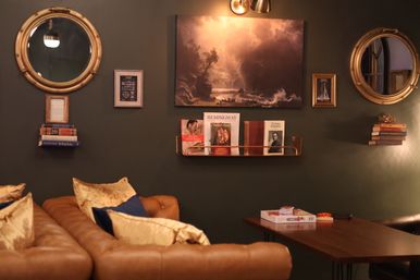 Cozy reading nook in a stylish lounge with a tufted tan leather sofa and gold pillows against a deep green wall, brass porthole mirrors, framed seascape art and shelves of books.