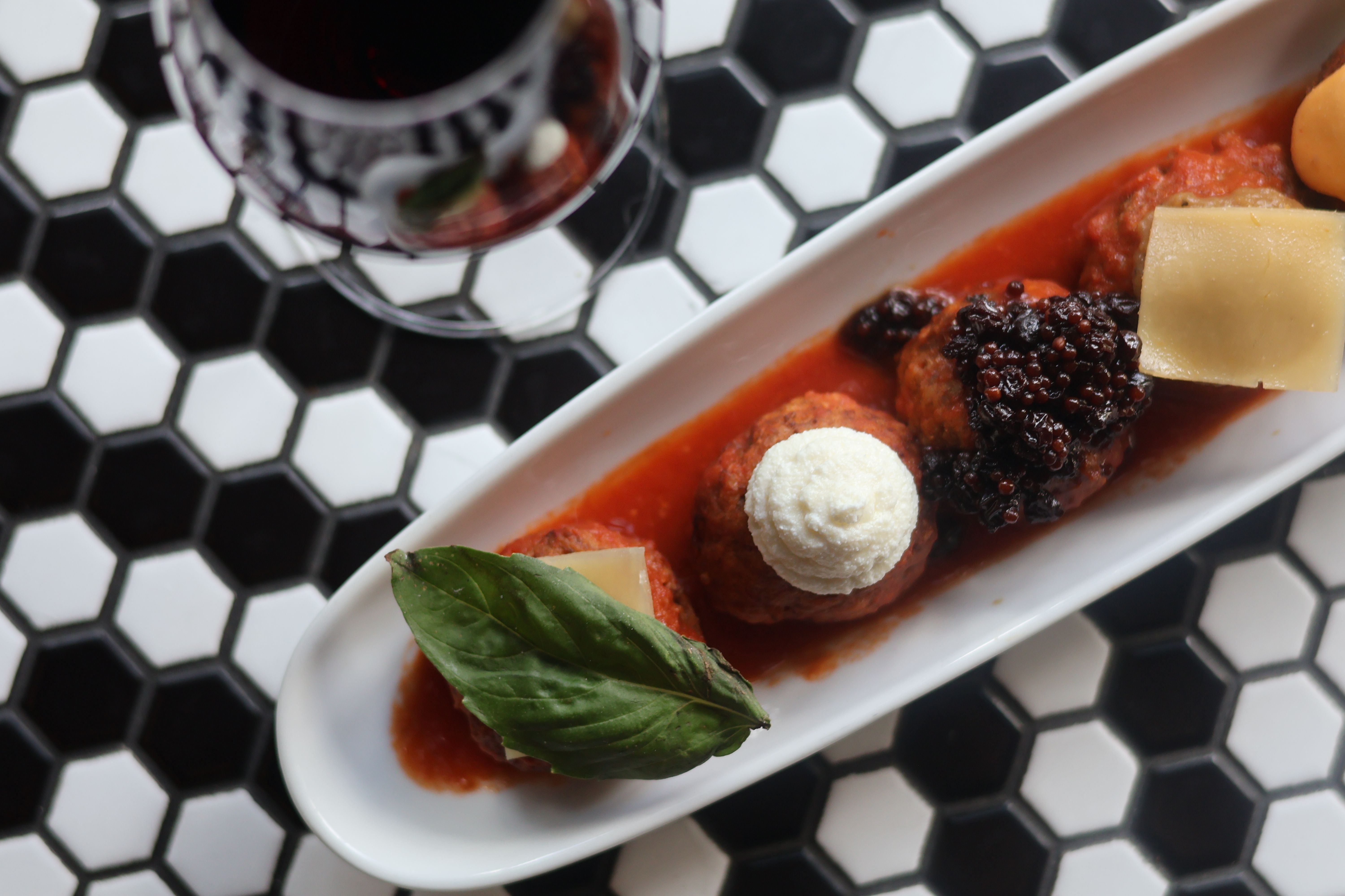 Top-down shot of Italian meatball appetizer in tomato sauce with a ricotta dollop, basil leaf, shaved parmesan and balsamic jam on a white dish beside a glass of red wine on black-and-white hex tile table