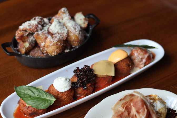 Assorted Italian-style meatball flight on a long white platter—each topped with basil, ricotta, relish, cheese slice and sauces—paired with a skillet of powdered-sugar fritters and a prosciutto-topped plate on a wooden table.