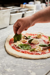 Hand placing fresh basil on a margherita pizza with tomato sauce, torn mozzarella and grated cheese on a floured countertop in a pizza kitchen
