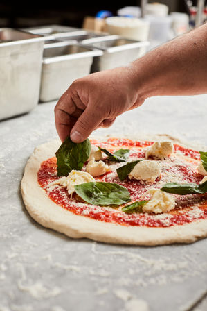 Hand placing fresh basil on a margherita pizza with tomato sauce, torn mozzarella and grated cheese on a floured countertop in a pizza kitchen