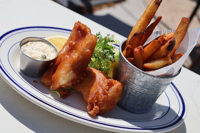 Golden beer-battered fish fillets with thick-cut fries in a small metal cup, tartar sauce in a metal ramekin, lemon wedge and parsley on a white blue-rimmed plate — classic fish and chips, outdoor dining.