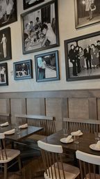 Cozy restaurant interior with wooden tables, spindle-back chairs and bench seating, neatly set plates and napkins, and a gallery wall of framed vintage black-and-white photos