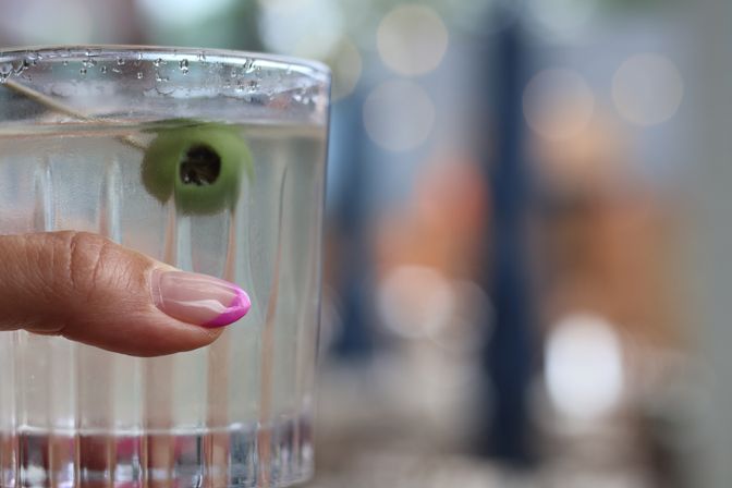 Close-up of a hand with pink French-tip manicure holding a ribbed cocktail glass with a green olive and condensation, soft bokeh lights blurred in the background.