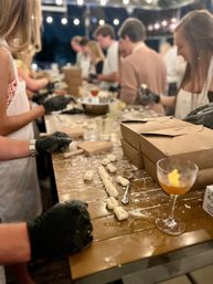Outdoor evening hands-on pasta-making class: people shaping gnocchi on a flour-dusted wooden table under string lights with takeout boxes and cocktails.