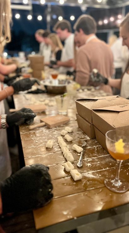Outdoor evening hands-on pasta-making class: people shaping gnocchi on a flour-dusted wooden table under string lights with takeout boxes and cocktails.