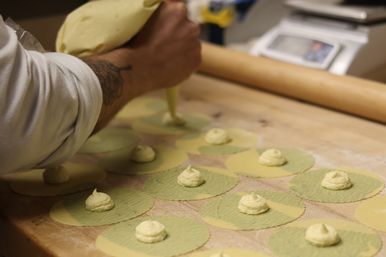 Hands using a piping bag to add creamy filling to green and yellow pasta rounds on a floured wooden board, with a rolling pin and scale in the background