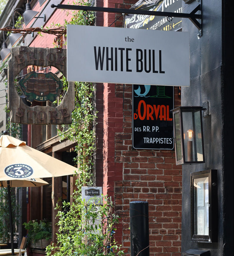 Hanging white pub sign above a red brick storefront with vintage wooden sign, lantern, climbing ivy and outdoor patio umbrella.