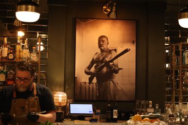 Dimly lit speakeasy-style bar with a bartender at the counter, mirrored shelves of bottles, and a large sepia portrait of a shirtless man holding a rifle on the wall.