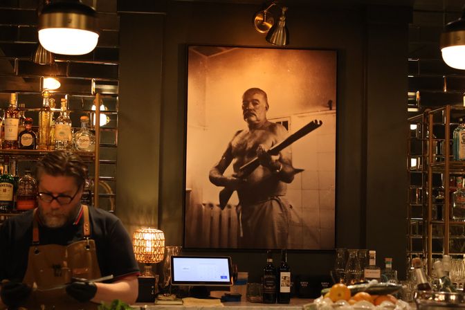 Dimly lit speakeasy-style bar with a bartender at the counter, mirrored shelves of bottles, and a large sepia portrait of a shirtless man holding a rifle on the wall.