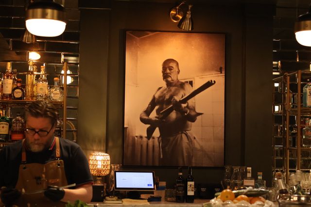 Dimly lit speakeasy-style bar with a bartender at the counter, mirrored shelves of bottles, and a large sepia portrait of a shirtless man holding a rifle on the wall.