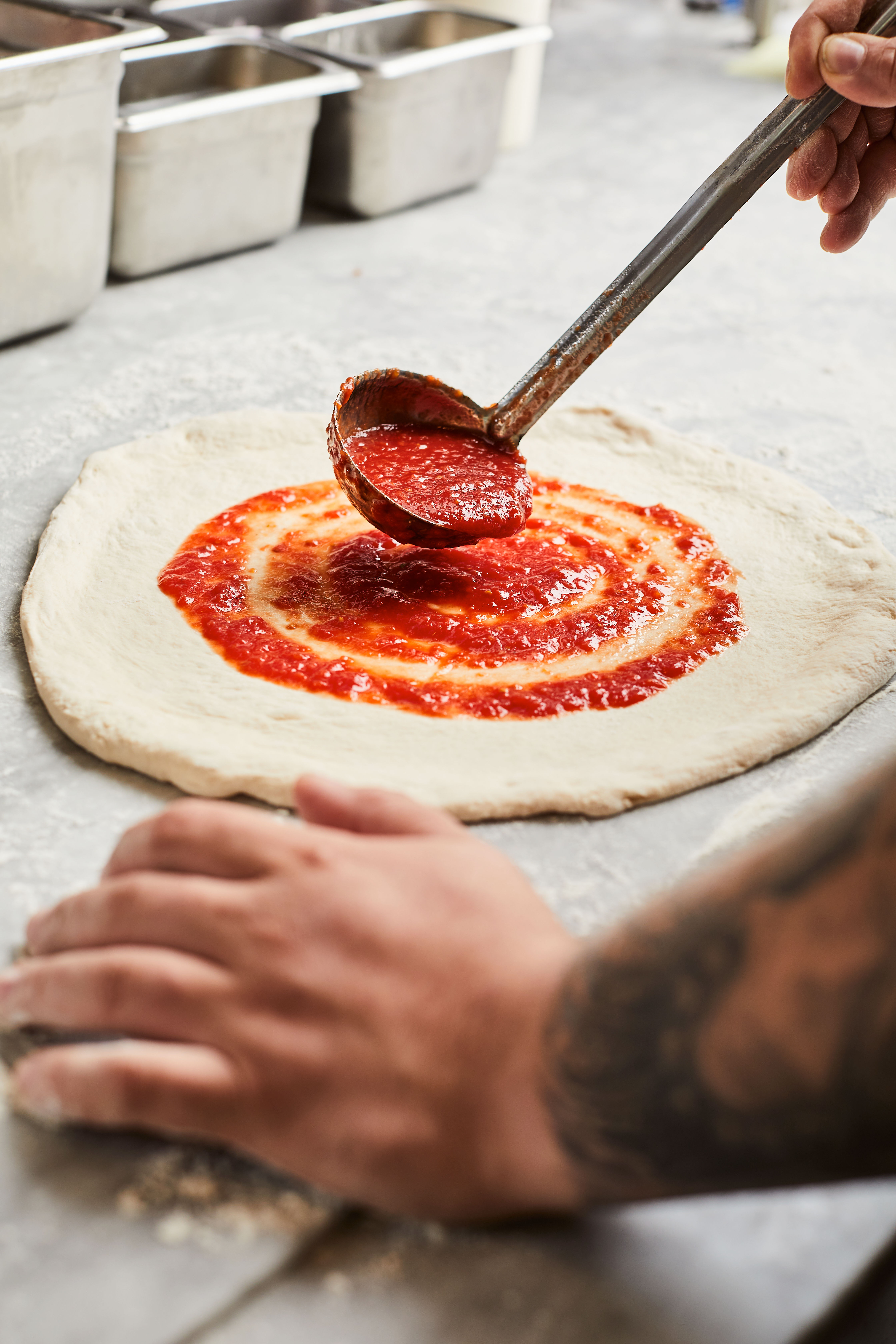 Close-up of a ladle spreading bright tomato sauce in a spiral over stretched pizza dough on a floured prep counter in a commercial kitchen