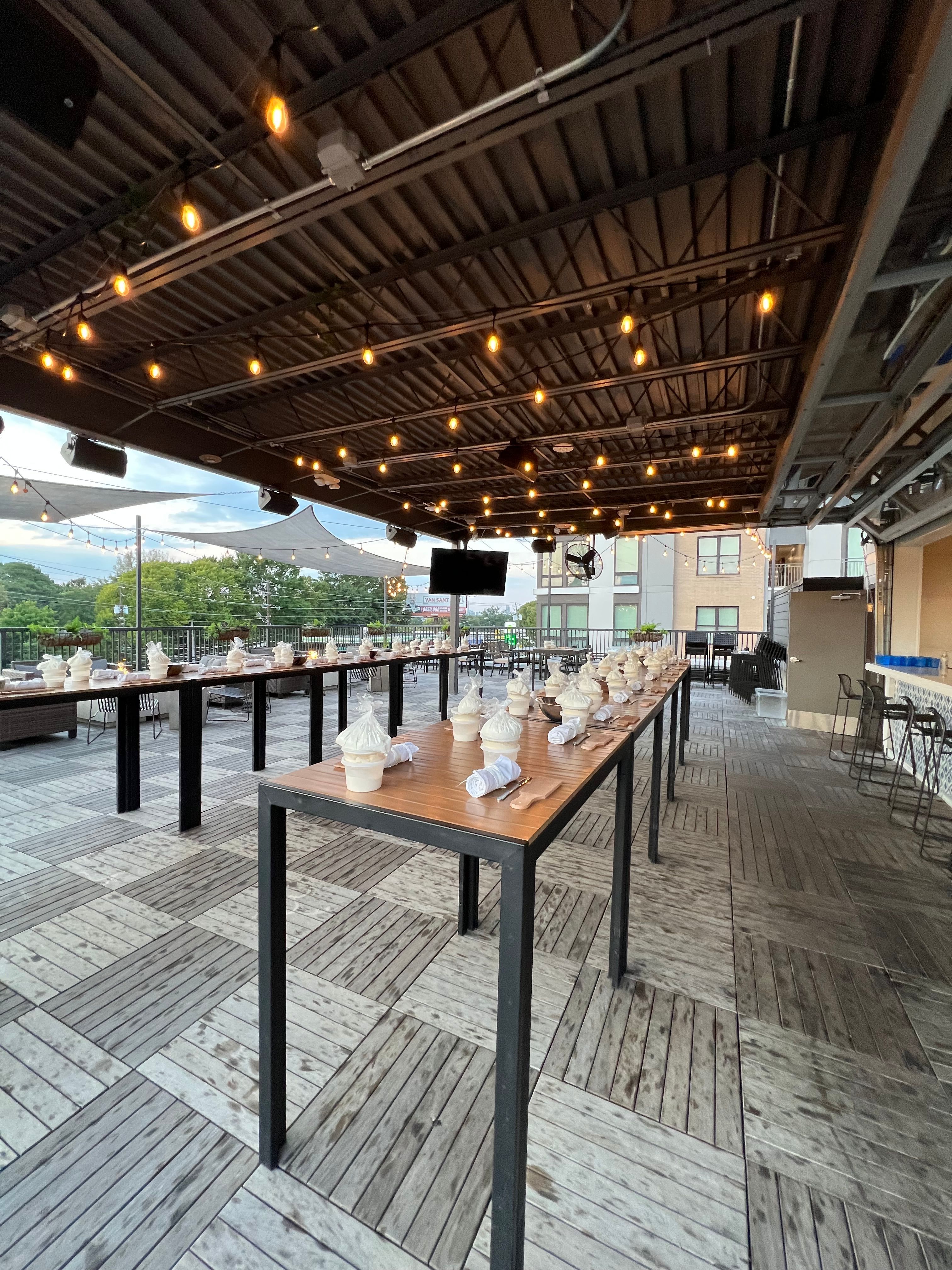 Rooftop patio at dusk with warm string lights, long wooden communal tables set with rolled napkins and ceramic cups, urban buildings and trees in background
