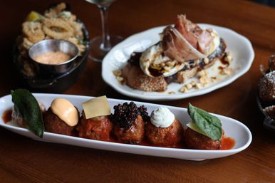 Five gourmet bite-size meatballs with varied sauces and garnishes in a white oval dish on a wooden table, framed by fried calamari with dipping sauce and prosciutto-topped crostini — small plates appetizer