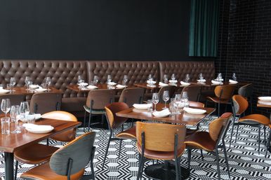 Modern restaurant interior with tufted brown leather banquette, wooden tables set with wine glasses and white napkins, curved wooden chairs and black-and-white geometric tile floor.