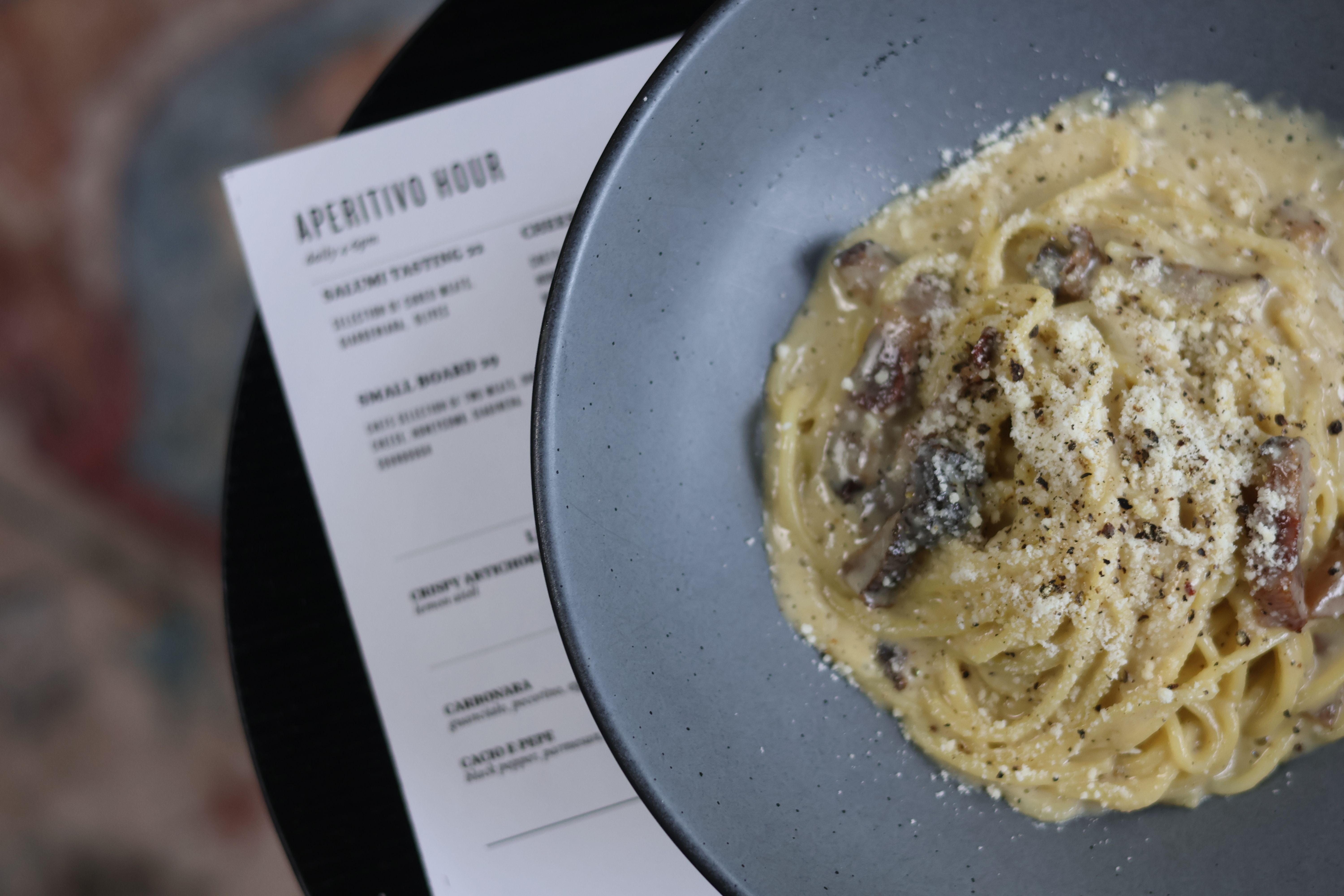 Close-up of creamy spaghetti carbonara sprinkled with grated cheese and cracked black pepper in a gray bowl, placed next to a printed "Aperitivo Hour" menu on a black table.