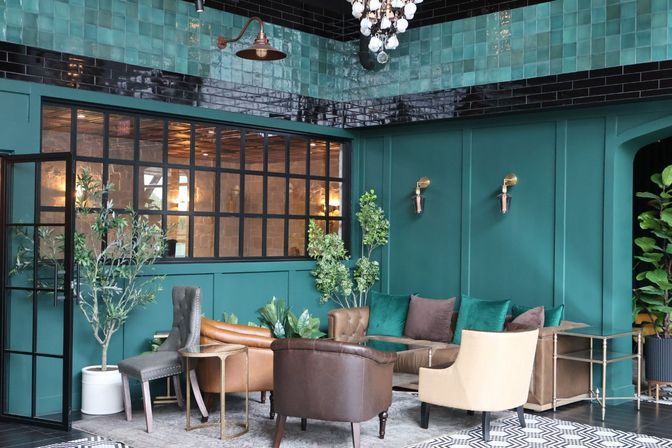 Cozy modern lounge interior with teal paneled walls and glossy black tile trim, leather and velvet seating with green pillows, patterned rug, potted plants, black-framed industrial glass partition and hanging chandelier — boutique urban lounge interior design.
