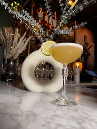 Golden cocktail in a coupe glass garnished with a lime wheel on a marble bar counter, with a modern white circular vase, eucalyptus stems, and backlit liquor bottles in an elegant bar setting.
