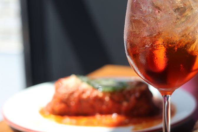 Close-up of a sunlit, chilled orange cocktail with ice in a stemmed glass, blurred plate of tomato-sauced pasta on a restaurant table in the background.