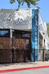 Urban white-brick oyster bar storefront with blue vertical signage, neon cursive logo, decorative metal patio screens and potted planters