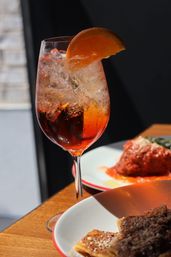 Sunlit icy orange spritz in a wine glass with an orange slice on a wooden table beside plates of pasta at an Italian restaurant.