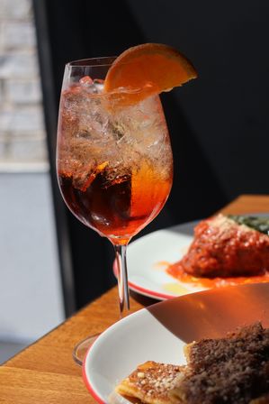 Sunlit icy orange spritz in a wine glass with an orange slice on a wooden table beside plates of pasta at an Italian restaurant.