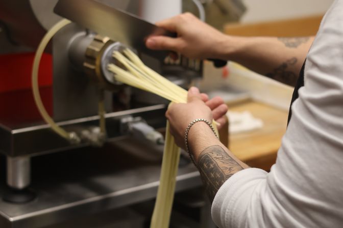 Chef's hands guiding fresh handmade pasta strands from a pasta extruder in a restaurant kitchen