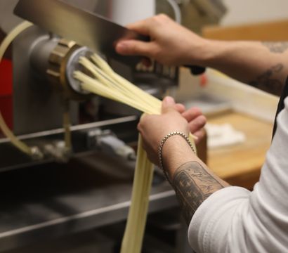 Chef's hands guiding fresh handmade pasta strands from a pasta extruder in a restaurant kitchen
