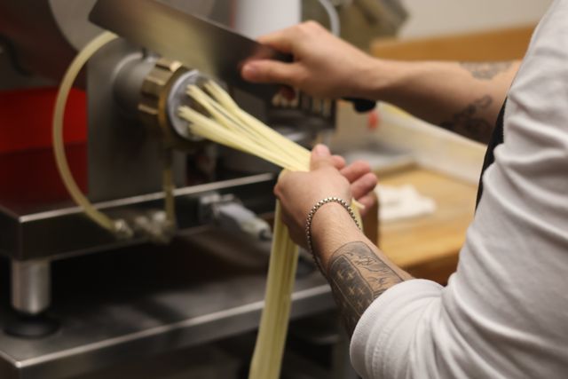 Chef's hands guiding fresh handmade pasta strands from a pasta extruder in a restaurant kitchen