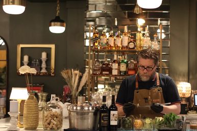 Bartender at an upscale cocktail bar preparing drinks at a marble counter with backlit shelves of spirits, wine bottles, citrus and herb garnishes, and a jar of corks.