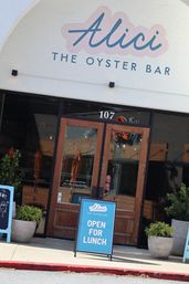 Street-level oyster bar entrance with white brick arched facade, wooden double doors numbered 107, cheerful teal A-frame 'Open for Lunch' sign on the sidewalk and potted greenery