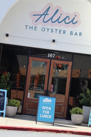 Street-level oyster bar entrance with white brick arched facade, wooden double doors numbered 107, cheerful teal A-frame 'Open for Lunch' sign on the sidewalk and potted greenery