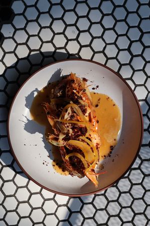 Gourmet grilled prawns topped with lemon peel and crunchy spice crumbs in a glossy orange-butter sauce on a white plate, photographed on black-and-white hexagon tile table