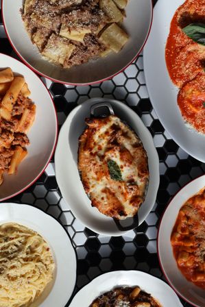 Top-down view of assorted Italian pasta dishes on black-and-white hex tile table: cheesy baked eggplant parmesan with basil in the center, surrounded by rigatoni, creamy cacio e pepe and several tomato-based pastas.