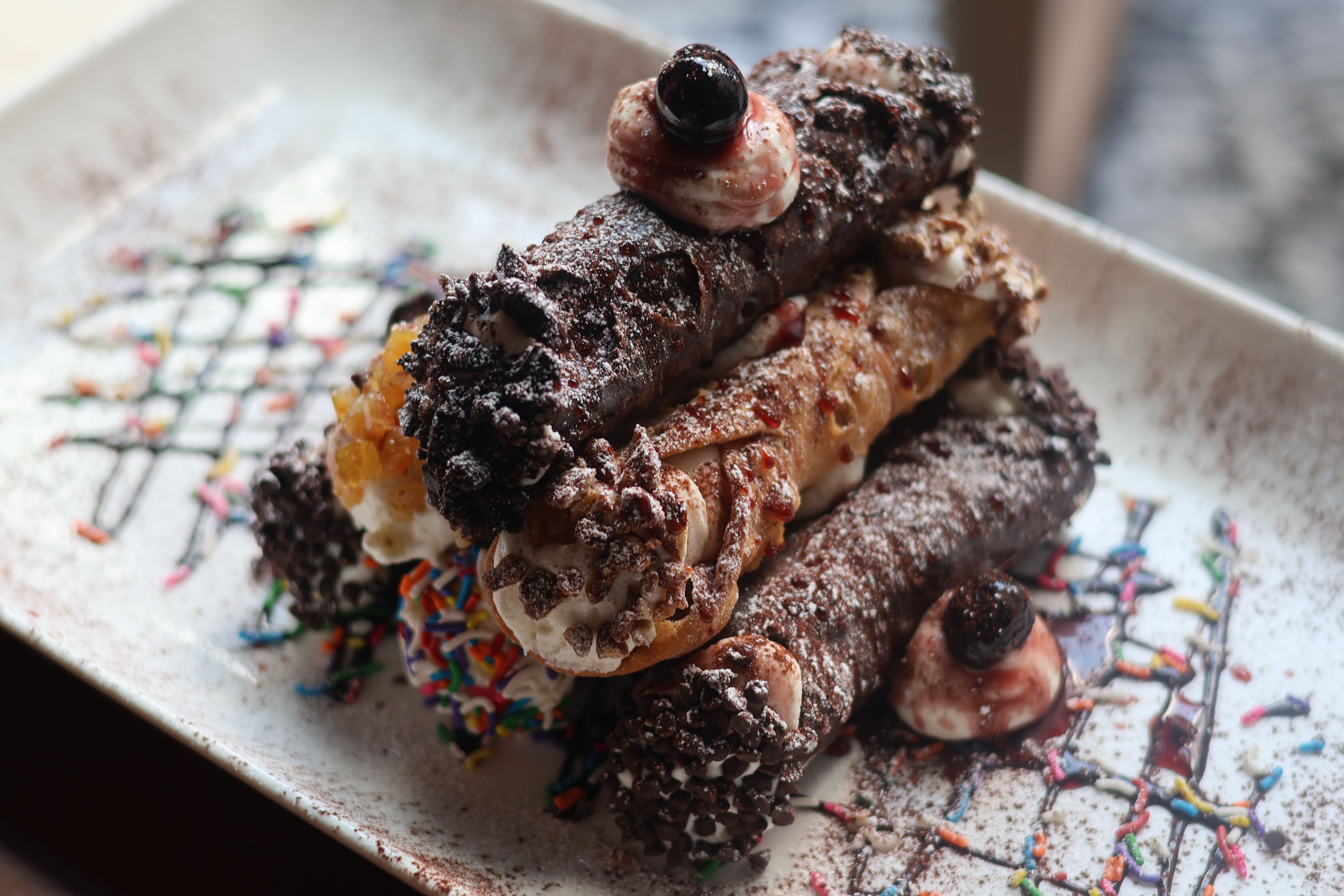 Close-up of stacked chocolate-dipped cannoli with creamy filling, chocolate chips, powdered sugar and rainbow sprinkles on a decorative dessert plate