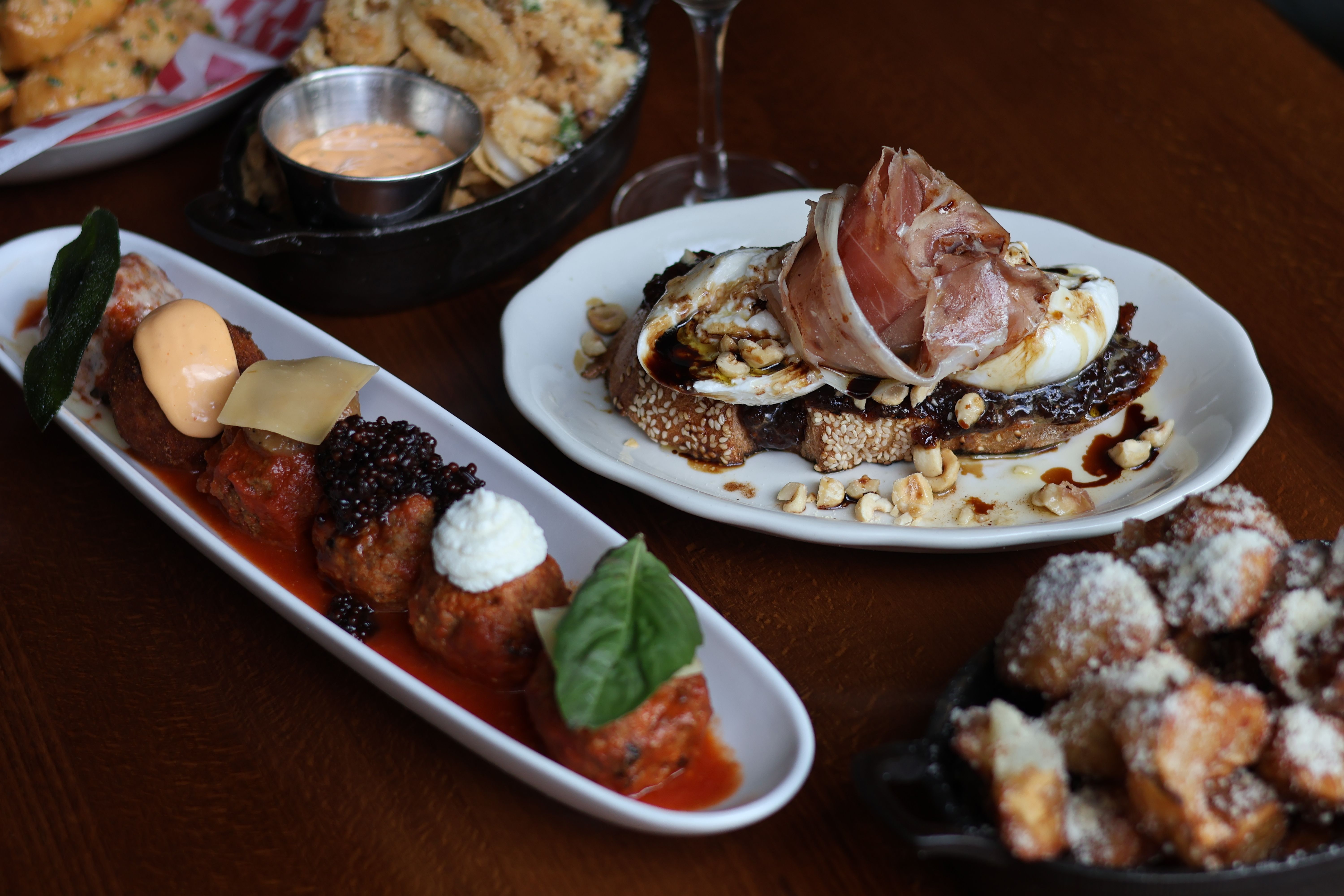 Assorted Italian small plates on a wooden table: sesame toast topped with burrata, prosciutto and balsamic glaze with chopped nuts, a long platter of sauced meatballs with different cheeses and basil, a skillet of fried dough bites dusted with grated cheese, and a small pan of crispy onion rings with dipping sauce.