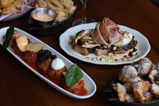 Assorted Italian small plates on a wooden table: sesame toast topped with burrata, prosciutto and balsamic glaze with chopped nuts, a long platter of sauced meatballs with different cheeses and basil, a skillet of fried dough bites dusted with grated cheese, and a small pan of crispy onion rings with dipping sauce.