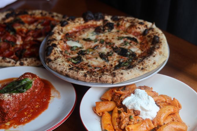 Wood-fired Neapolitan pizza with charred crust, rigatoni in rich tomato-meat sauce topped with creamy ricotta, and a saucy meatball in marinara with basil served on white plates on a wooden table.