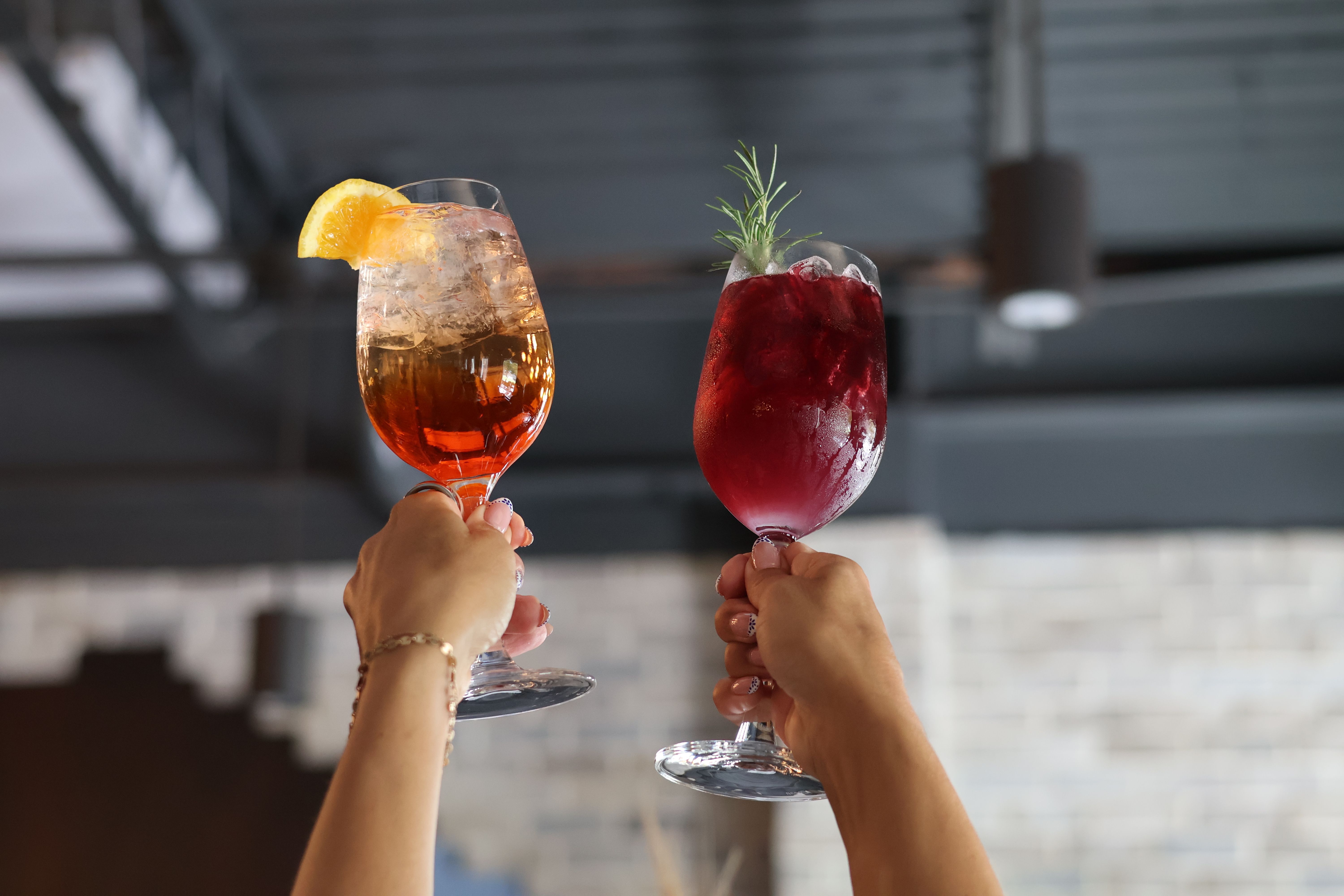 Two hands toasting colorful cocktails in wine glasses — amber drink with ice and an orange slice and a deep red drink garnished with rosemary — raised against a modern indoor bar ceiling.