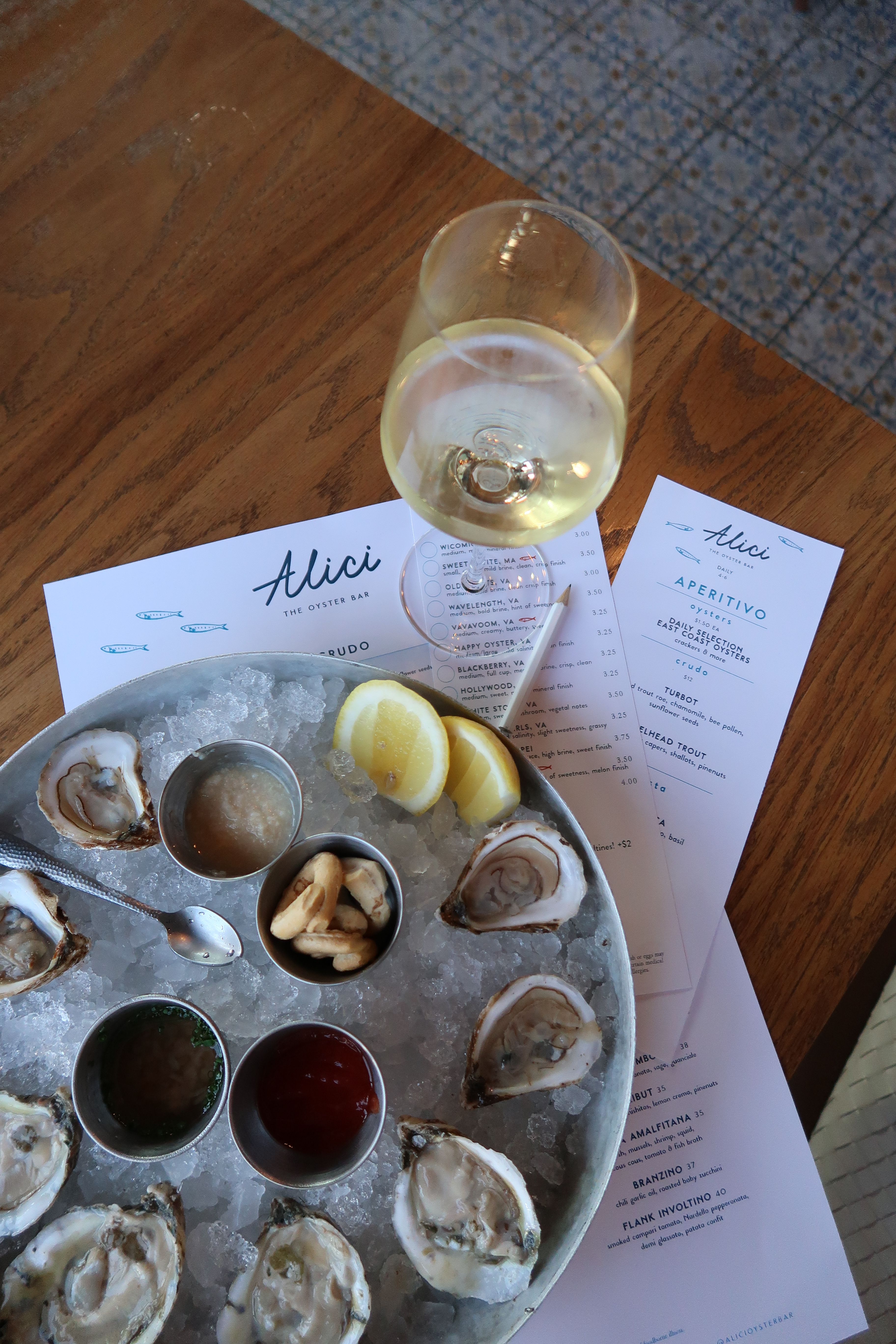Top-down view of a chilled raw oyster platter on crushed ice with lemon wedges, sauce cups and crackers, paired with a glass of white wine and restaurant menus on a wooden table