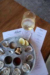 Top-down view of a chilled raw oyster platter on crushed ice with lemon wedges, sauce cups and crackers, paired with a glass of white wine and restaurant menus on a wooden table