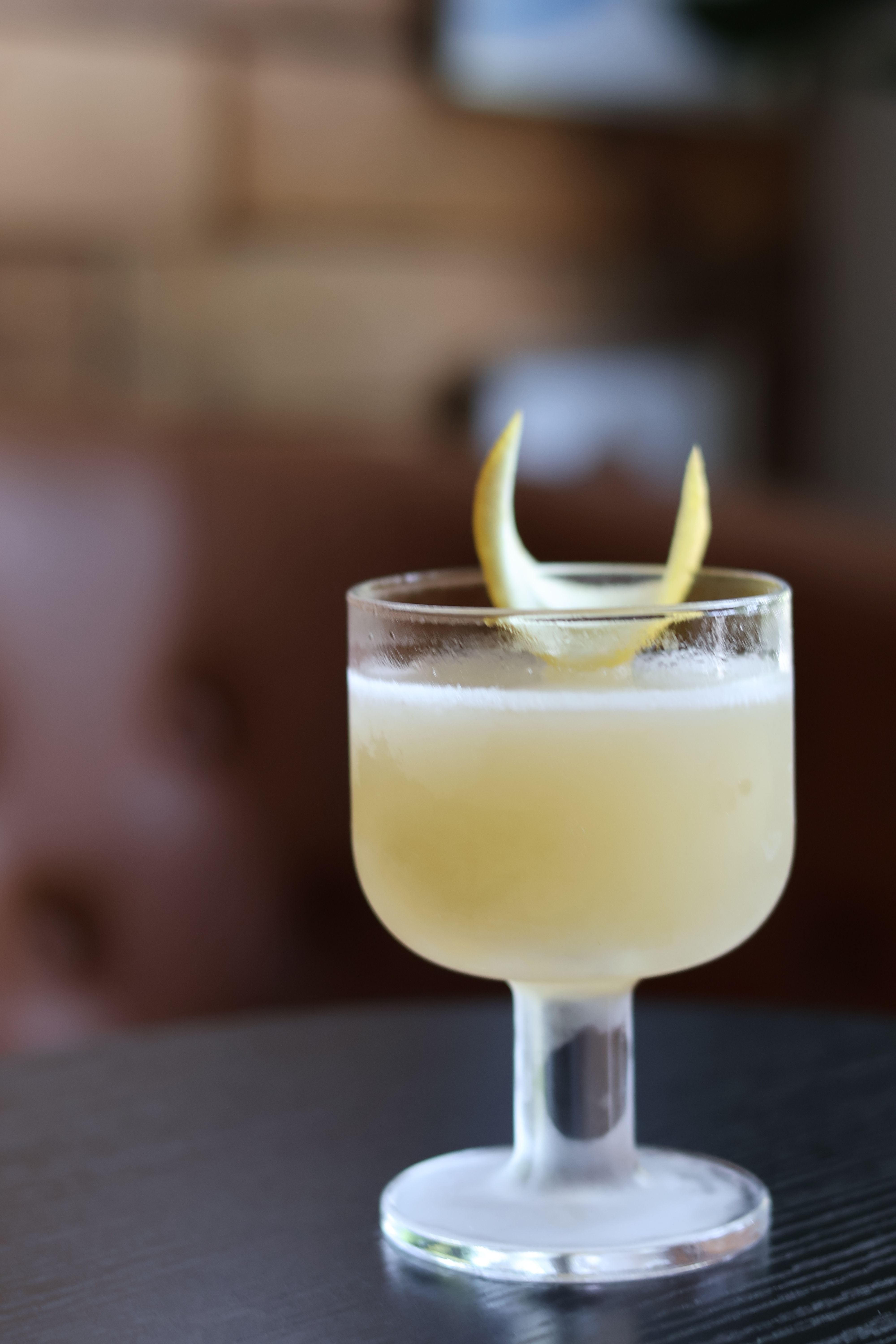 Frosted stemmed cocktail glass with a pale yellow citrus drink and lemon-twist garnish on a dark table, blurred lounge background