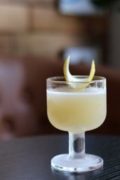 Frosted stemmed cocktail glass with a pale yellow citrus drink and lemon-twist garnish on a dark table, blurred lounge background