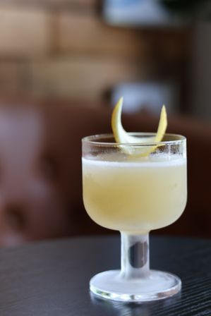 Frosted stemmed cocktail glass with a pale yellow citrus drink and lemon-twist garnish on a dark table, blurred lounge background