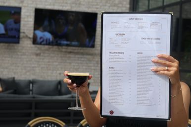 Outdoor patio restaurant scene: a person holds a menu titled "Lunch Cocktails" in one hand while holding a dark, creamy-topped cocktail in a coupe glass with the other, set against a brick wall with lounge seating and wall-mounted TVs.