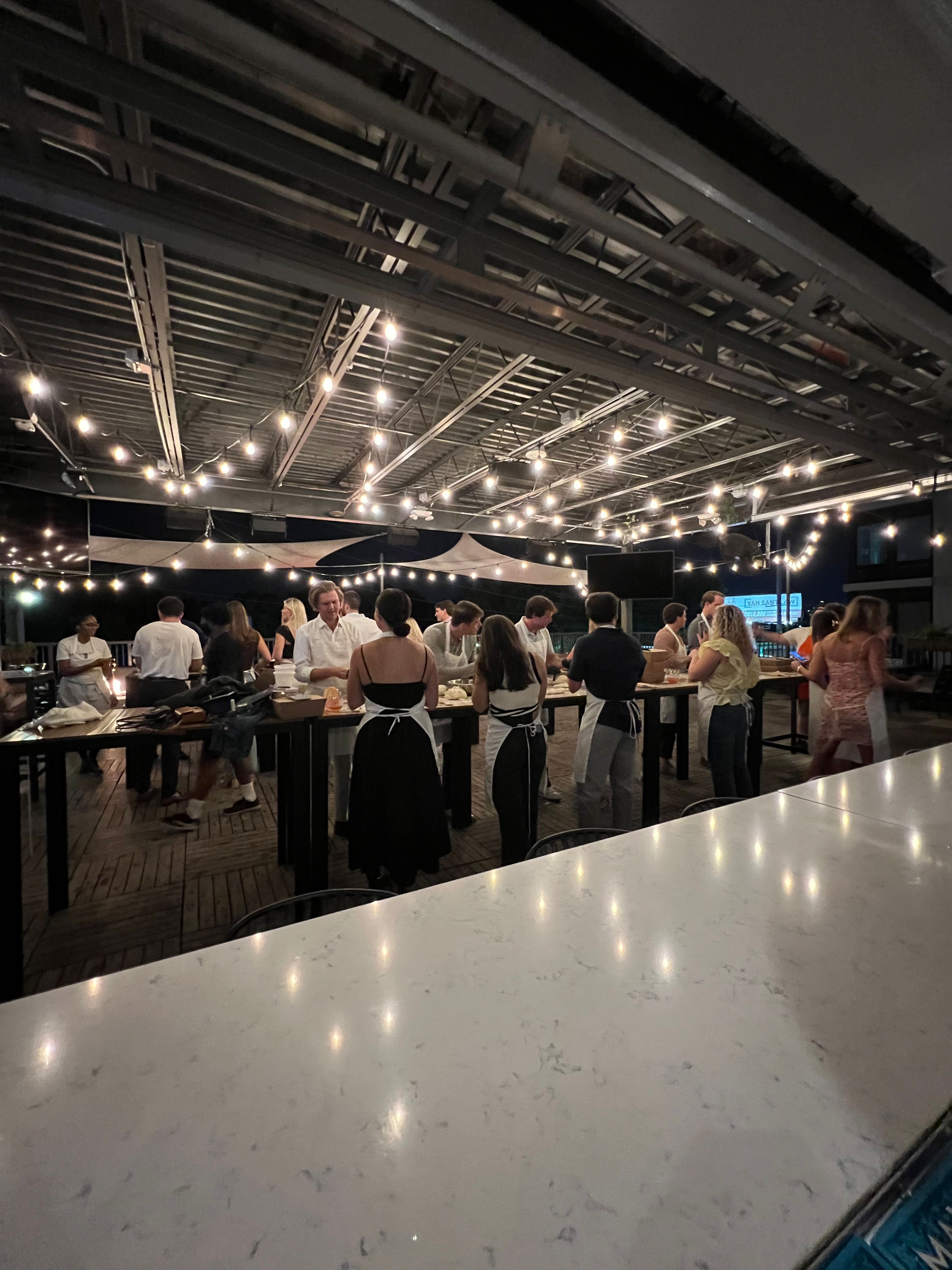 Rooftop patio at night with hanging string lights and a group of people in aprons gathered around long high tables for a communal cooking class/outdoor dining event